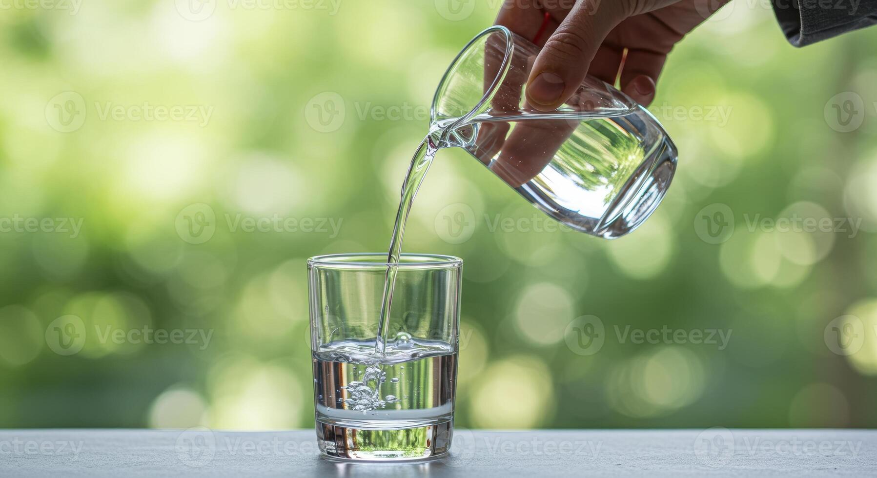 Hand pours clear liquid from glass pitcher into tumbler. Refreshment concept with blurred green foliage in background. Hydration and wellness theme. photo