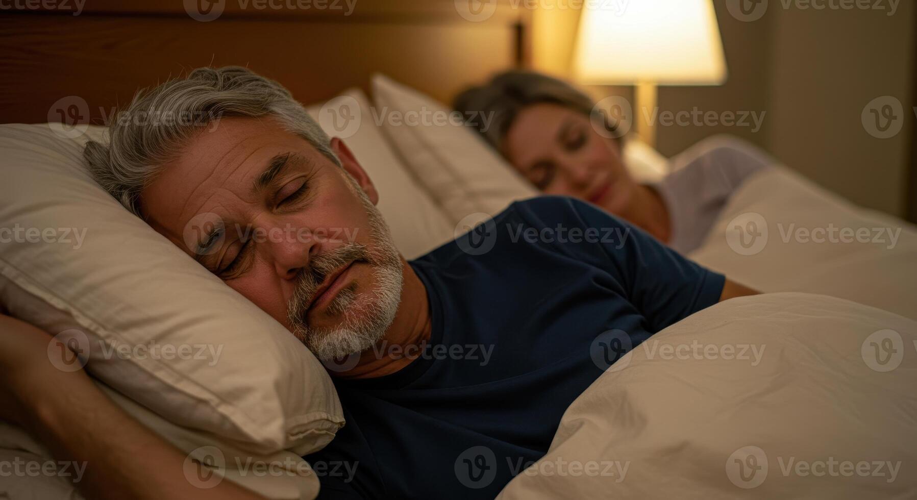 Mature couple sleeping in bed at night. Man with gray hair and beard asleep on pillow next to woman in soft focus. Bedroom illuminated by lamp. photo