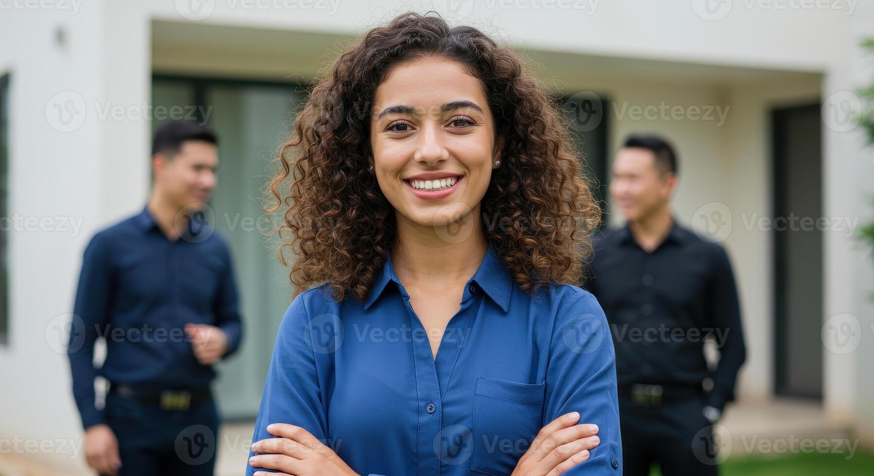 Smiling woman with curly hair standing with arms crossed in front of house with two men in background. Real estate agent concept. Confident female professional. photo
