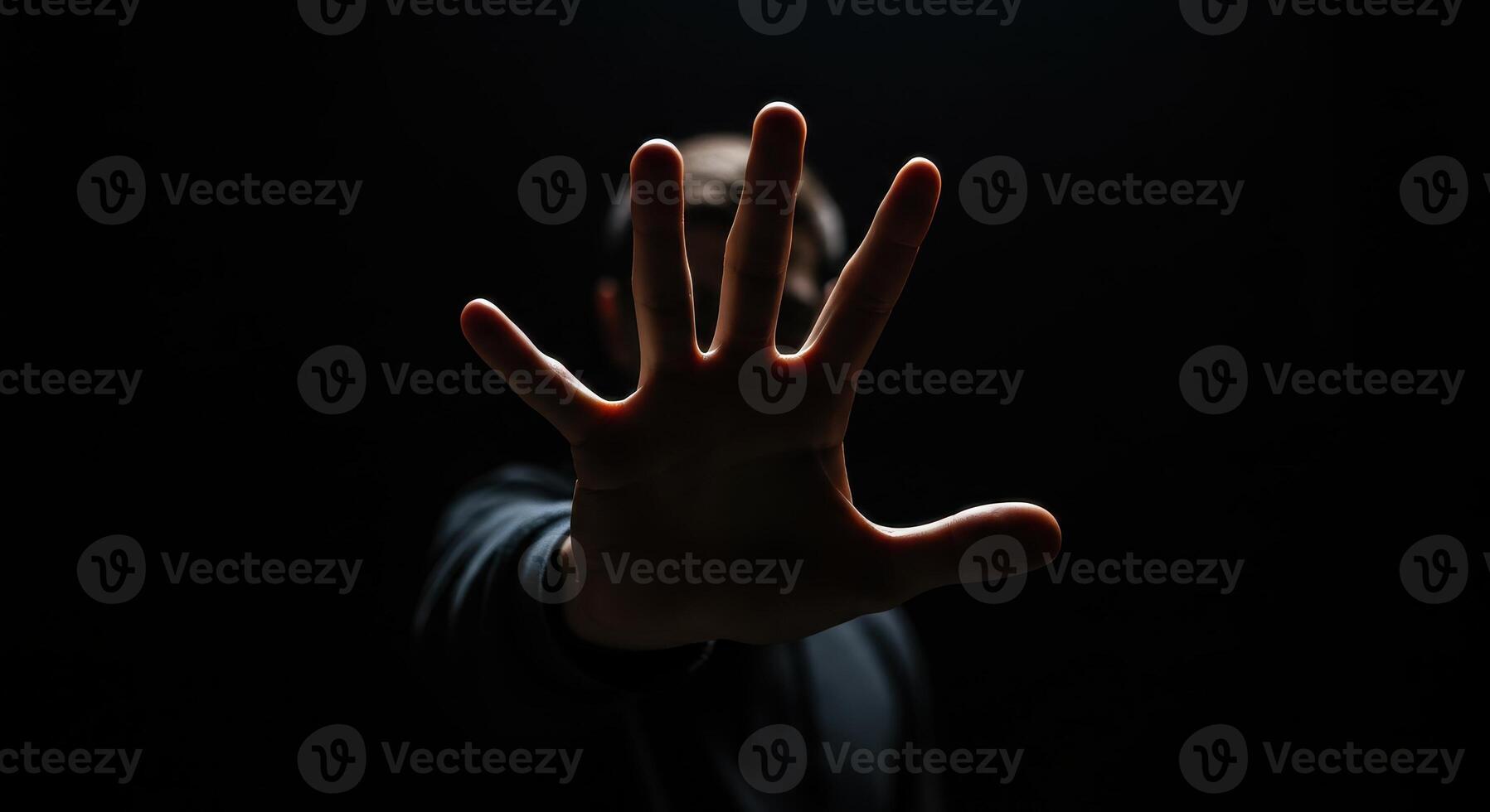 Man raises hand in front of face in dark setting, fingers spread. Gesture of stopping, blocking, or Vulcan salute concept on black background. photo