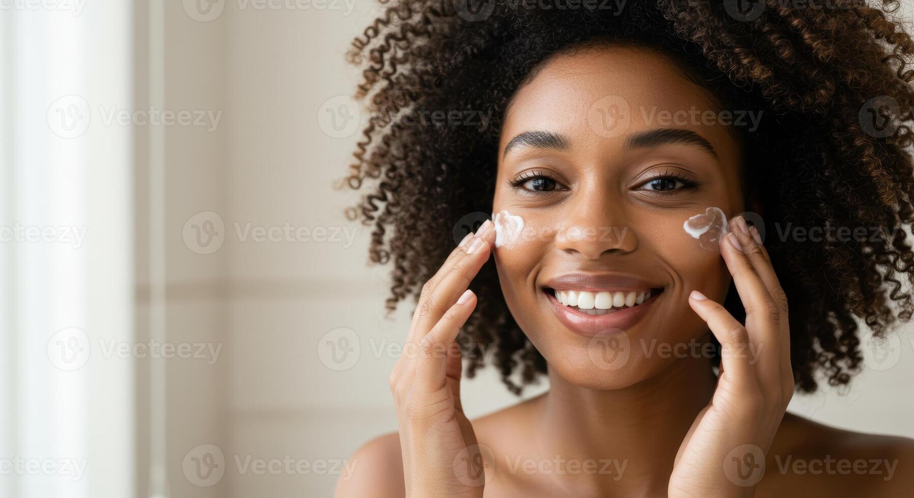 Smiling African American woman applies moisturizer to her face. Skincare routine with natural curly hair. Beauty treatment for healthy skin and radiant complexion. photo