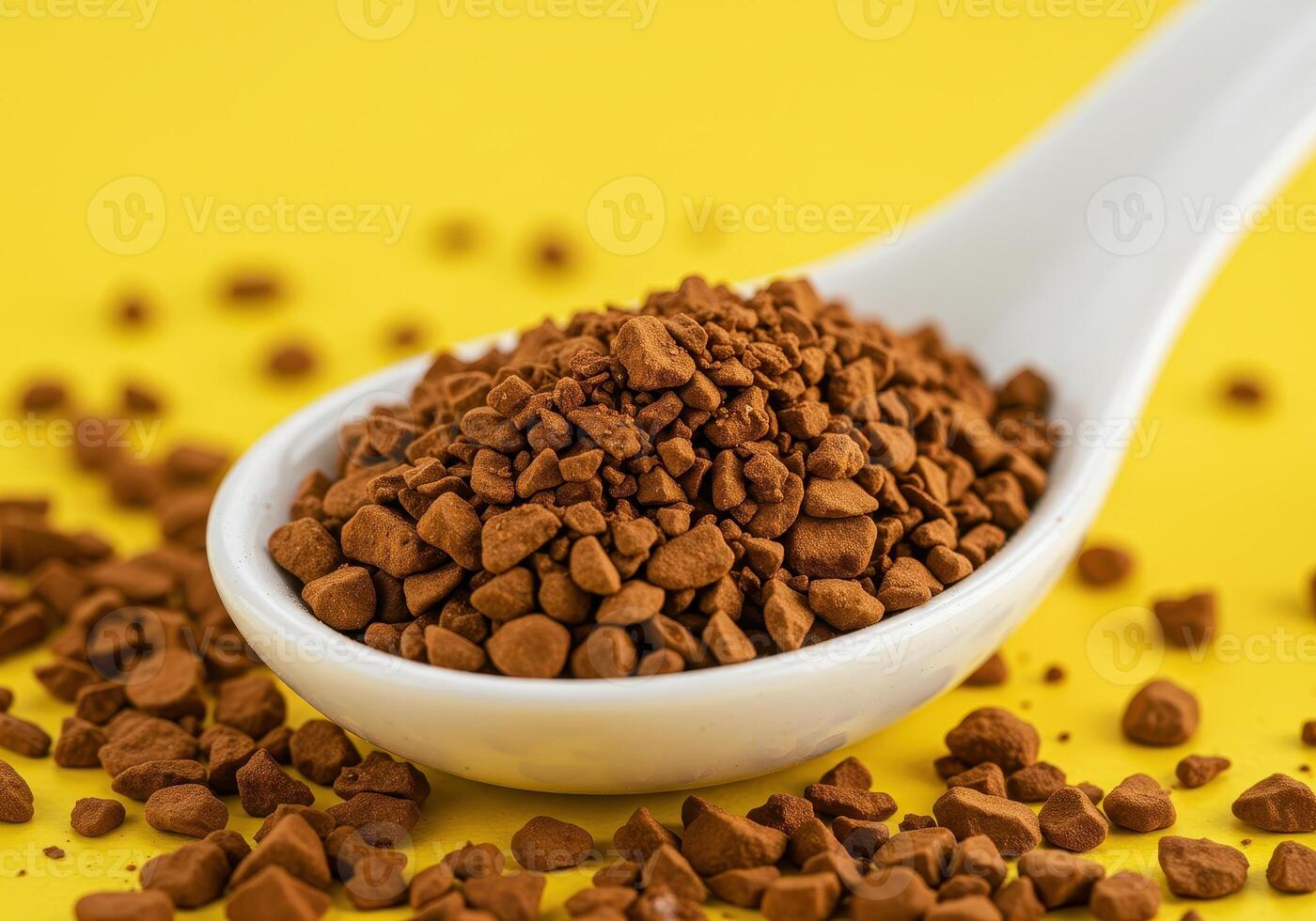Close up of instant coffee granules on a white ceramic spoon against a bright yellow backdrop. Coffee granules are also scattered around the spoon. photo