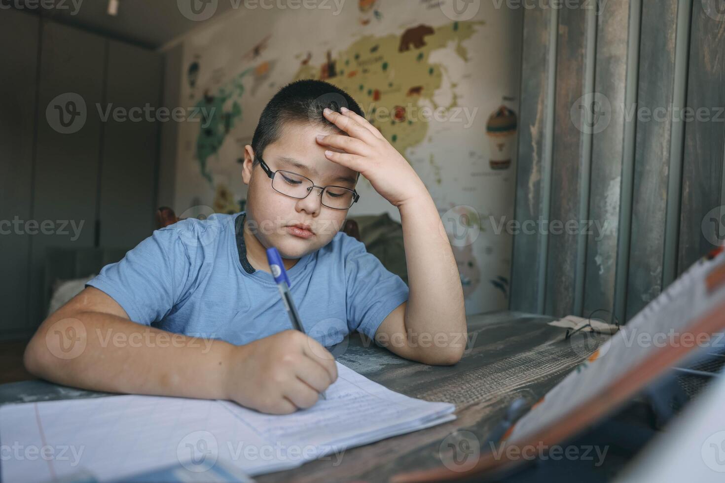 asian boy with glasses holding head in frustration, school task at home desk near world map wallpaper, concept of homeschooling, education service, tutoring support photo