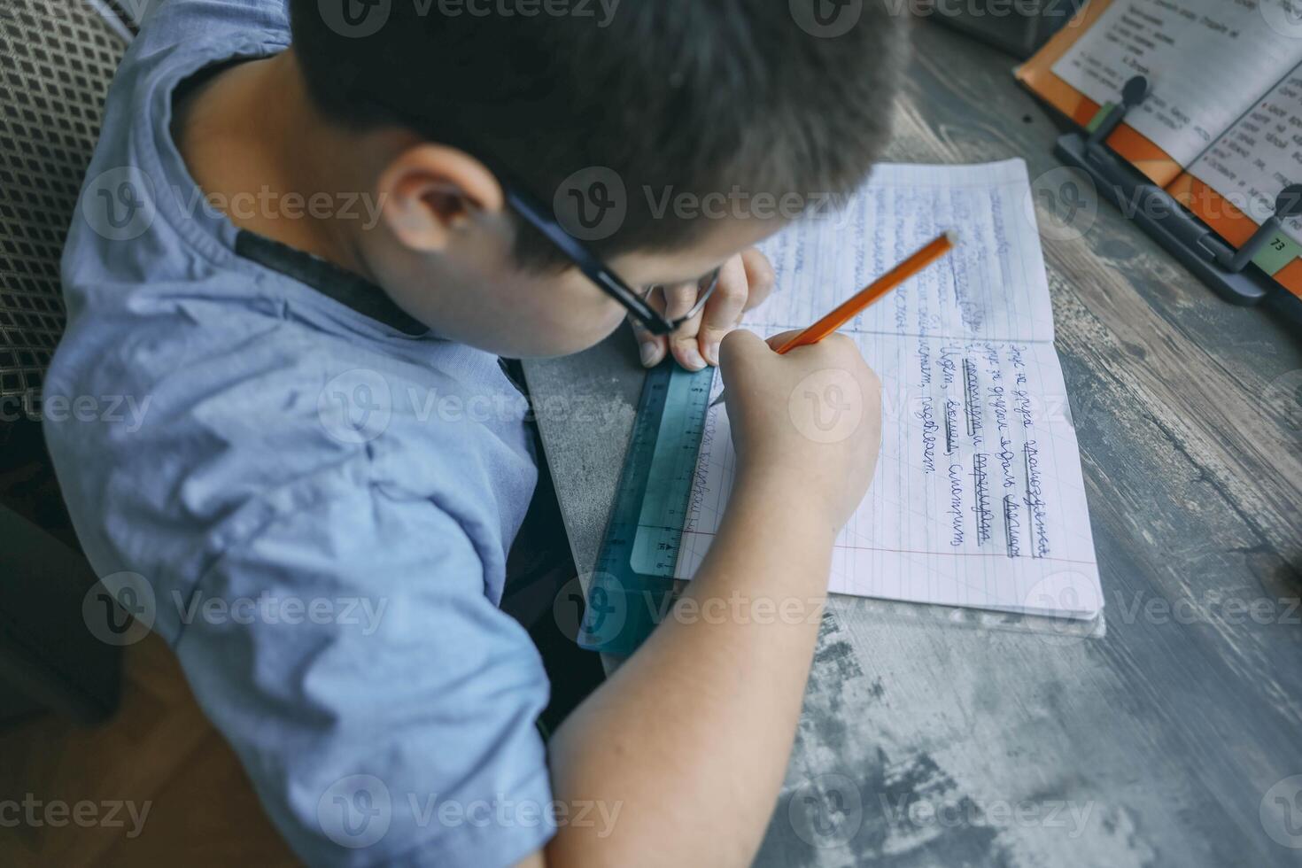 boy in blue shirt using ruler and pencil while doing homework, close up of child writing in notebook, wooden desk with textbook photo