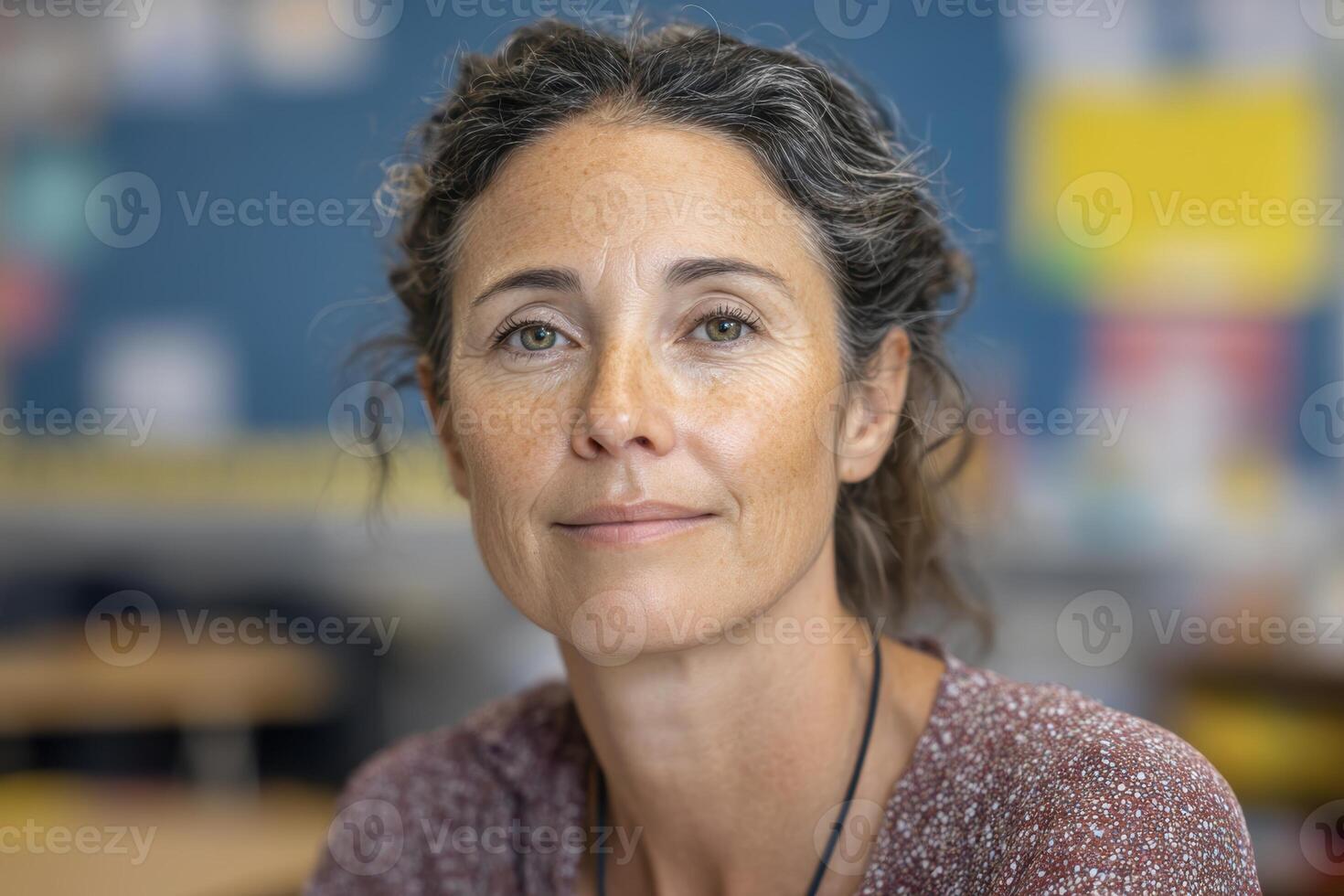 A woman with curly hair, wearing a patterned shirt, looks into the camera with a calm expression. The background is slightly blurred, suggesting an indoor setting such as an office or classroom photo