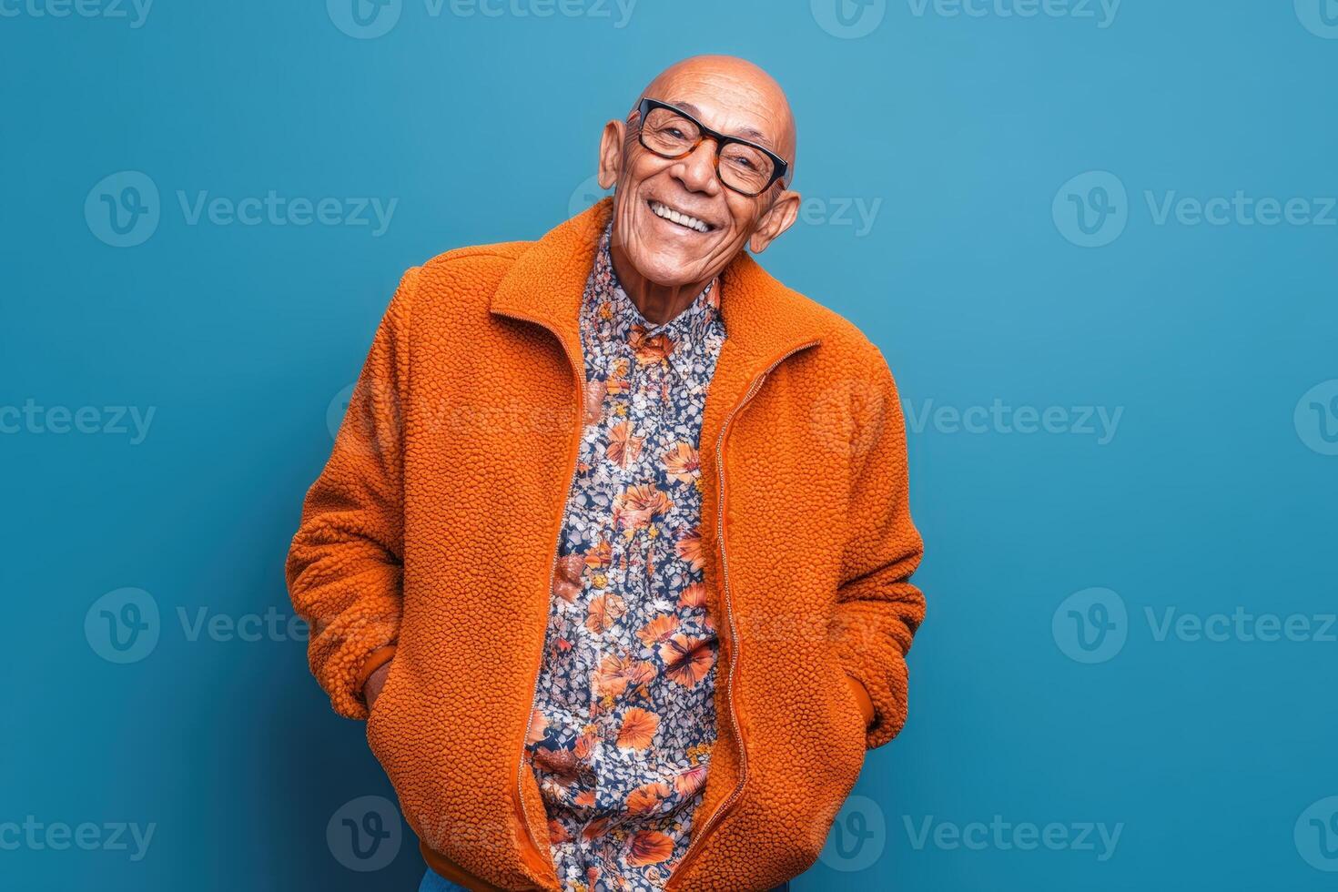 Elderly man in glasses smiling warmly, wearing an orange jacket and floral-patterned shirt. He stands with hands in pockets against a blue wall, exuding confidence and joy photo
