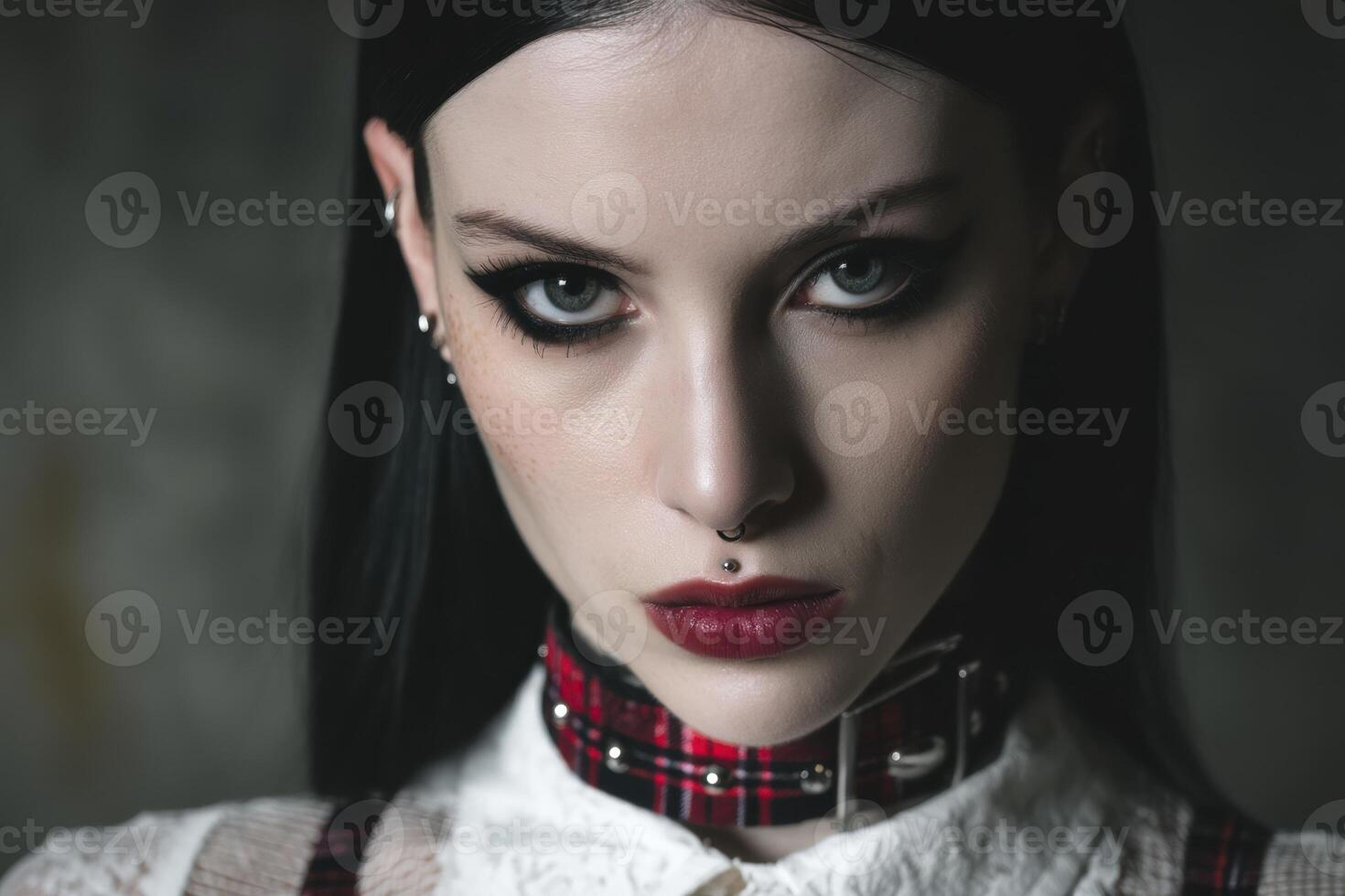 Dark-haired person with striking eye makeup, red lips, and facial piercings. They wear a plaid choker over a white collared top. Intense gaze. Earring studs visible photo