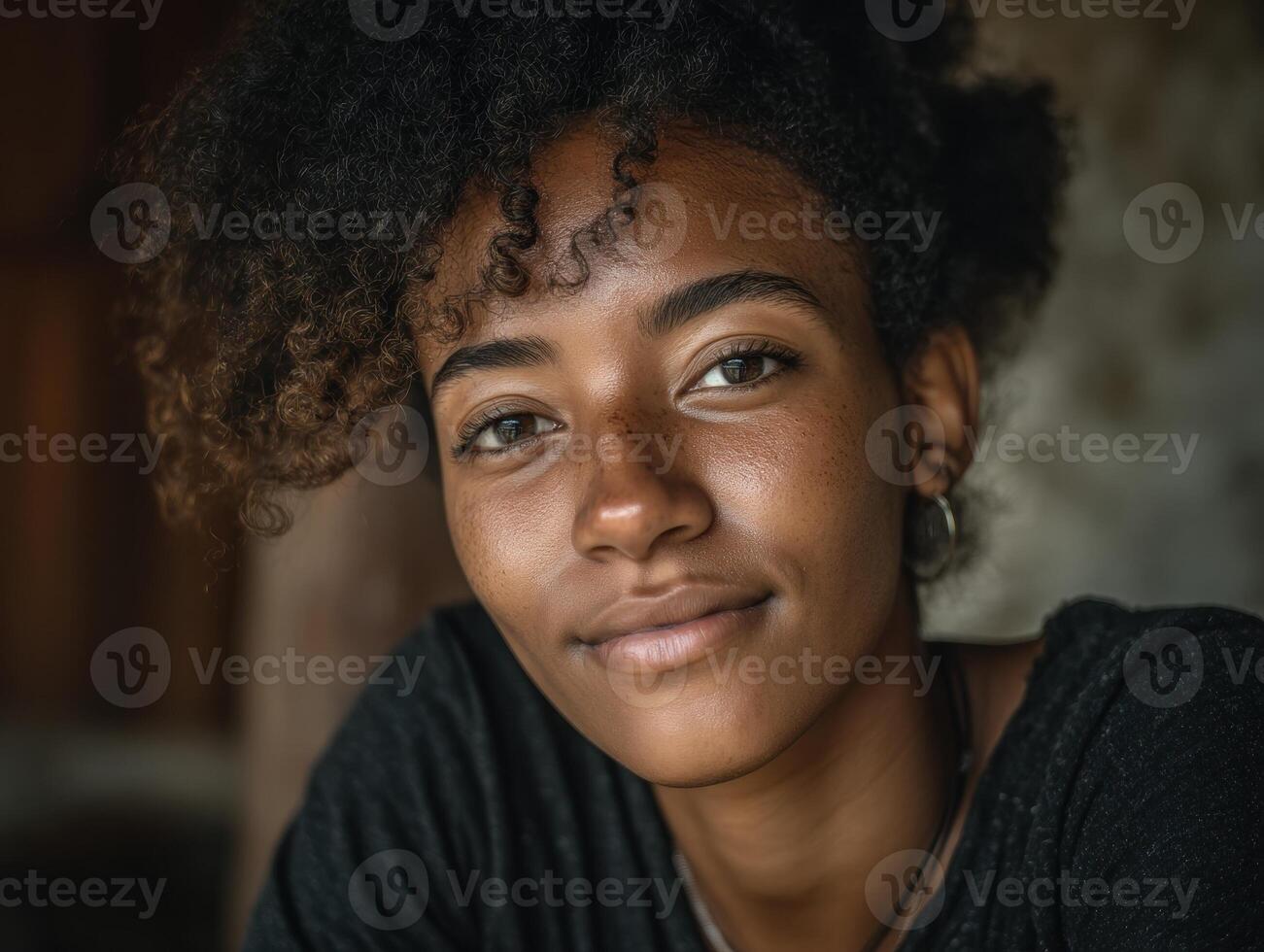 Young person with curly hair smiles warmly, wearing a dark top and earrings. They are standing in a softly lit environment with natural shadows enhancing facial features. Expression is serene photo