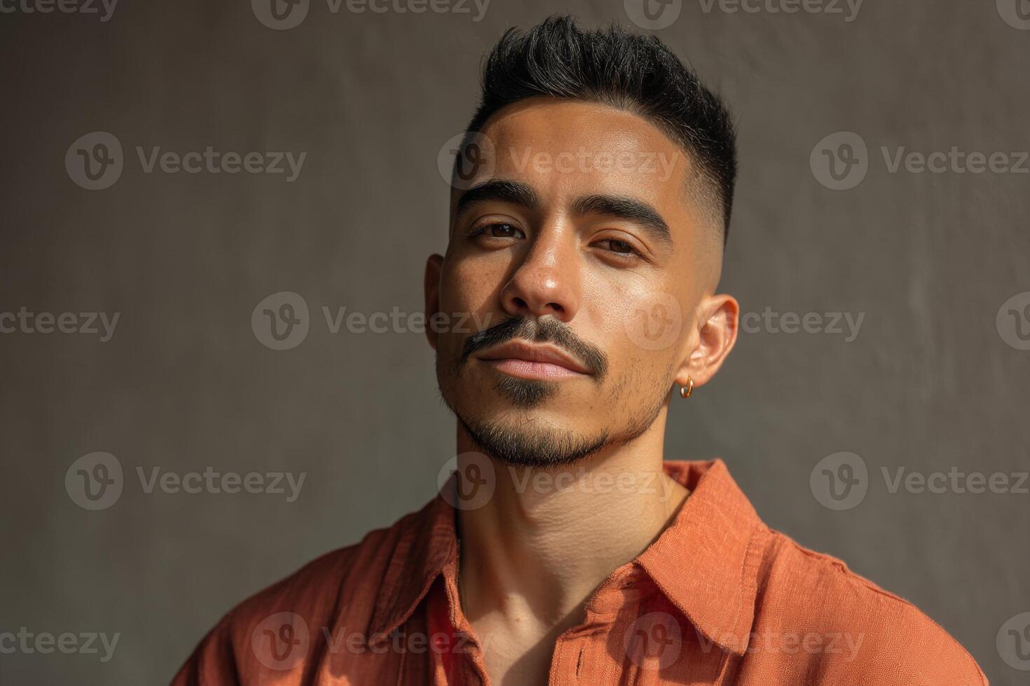 Portrait of a man with short dark hair, wearing an orange shirt. He has a beard, mustache, and an earring. Light falls on his face, creating a soft shadow on the right side photo