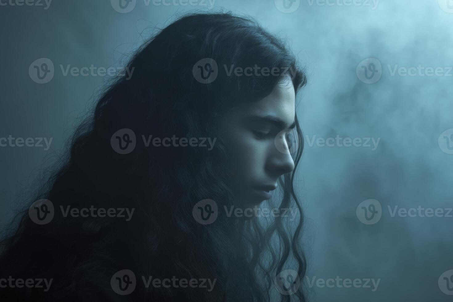 Profile view of a young person with long curly hair and closed eyes, surrounded by soft fog. The lighting is moody, emphasizing the serene and contemplative expression photo