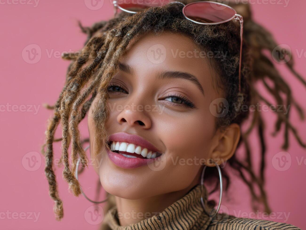 Smiling woman with dreadlocks and large hoop earrings, wearing turtleneck sweater and sunglasses on head. Pink background accentuates her bright smile and natural beauty. Close-up shot photo