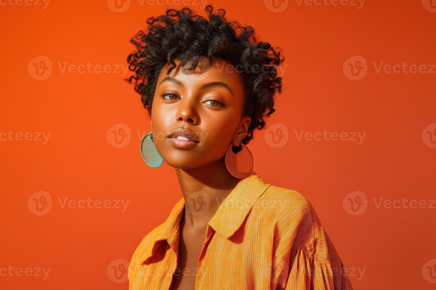 Curly-haired person with hoop earrings wearing an orange shirt, looking confidently at the camera. The vibrant backdrop complements the warm tones of the outfit and accessories photo