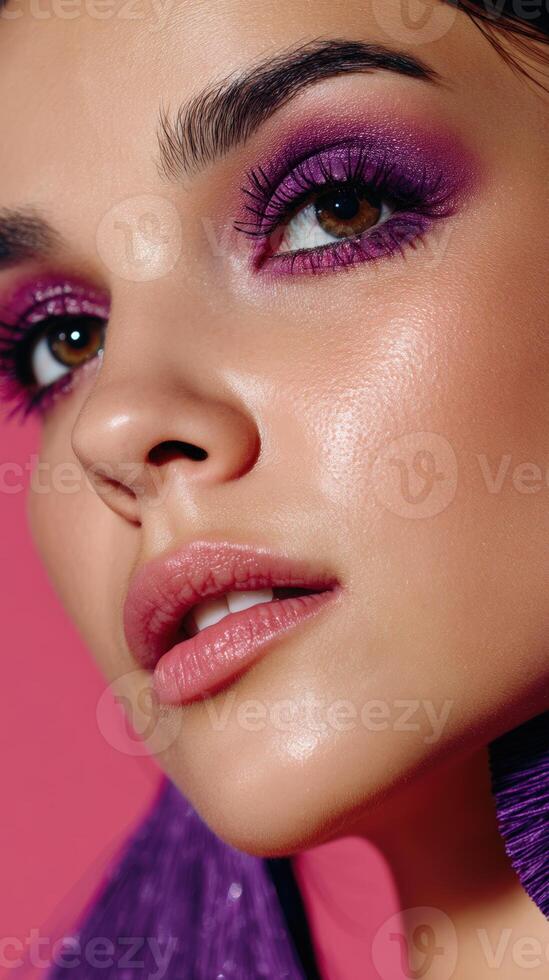 Close-up of a woman's face wearing vibrant purple eyeshadow and long lashes, with glossy lips and a smooth complexion, looking upwards photo