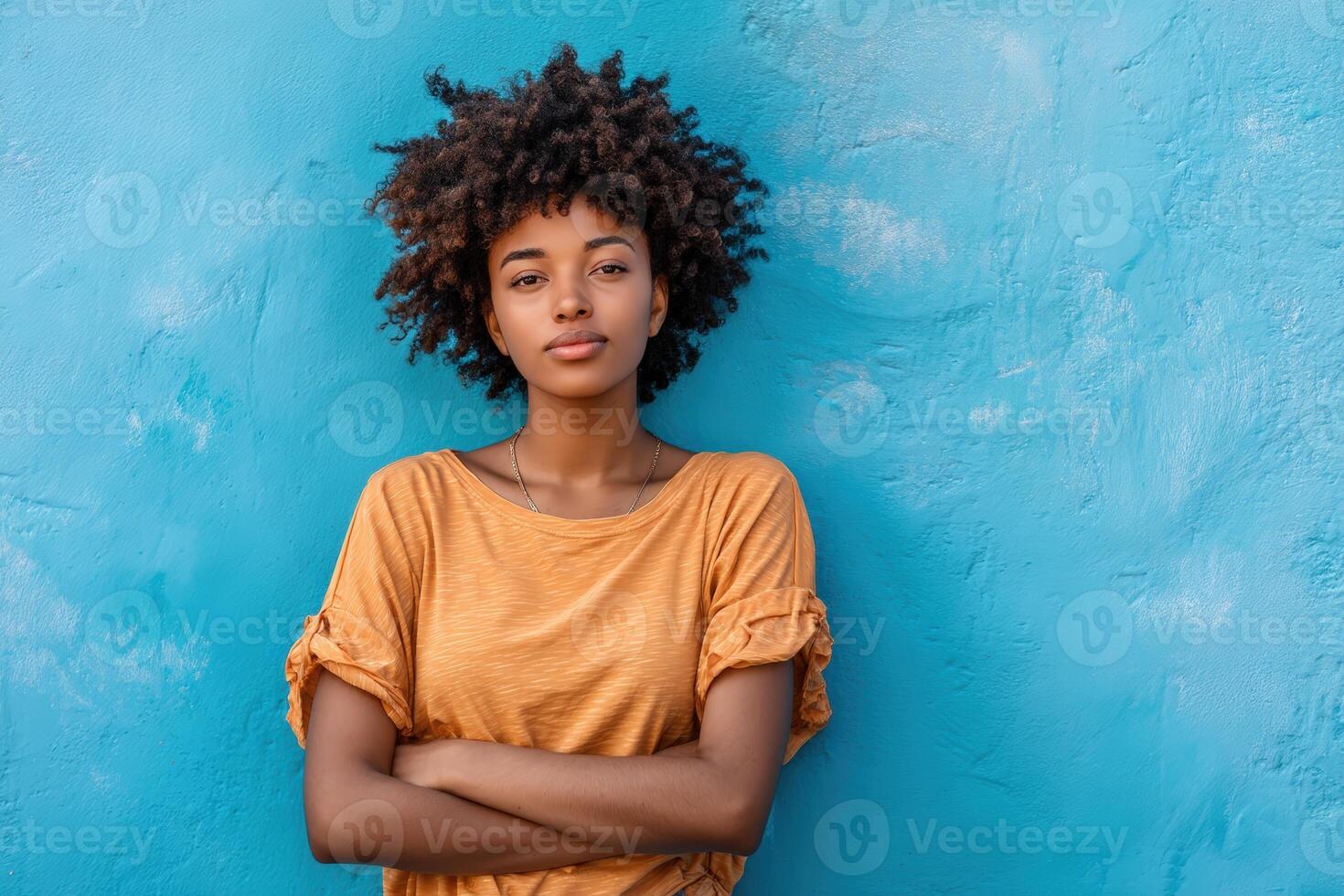 Young person with curly hair, wearing a yellow shirt, stands confidently with crossed arms. Bright blue wall serves as a vivid backdrop, highlighting the relaxed and casual vibe photo