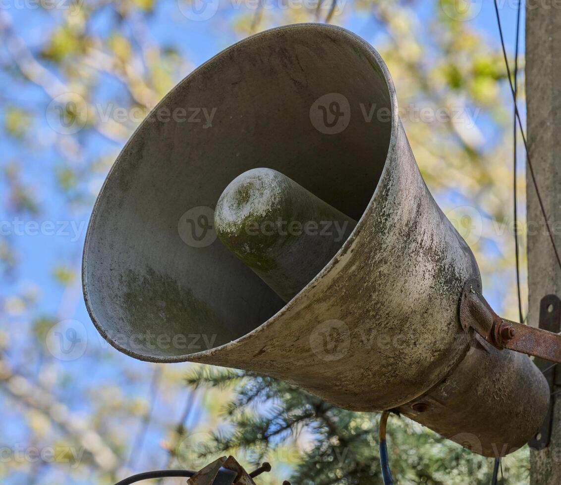 An old, dirty, grey loudspeaker is mounted on a pole. In the background, there is a blurred image of the sky and tree branches. photo