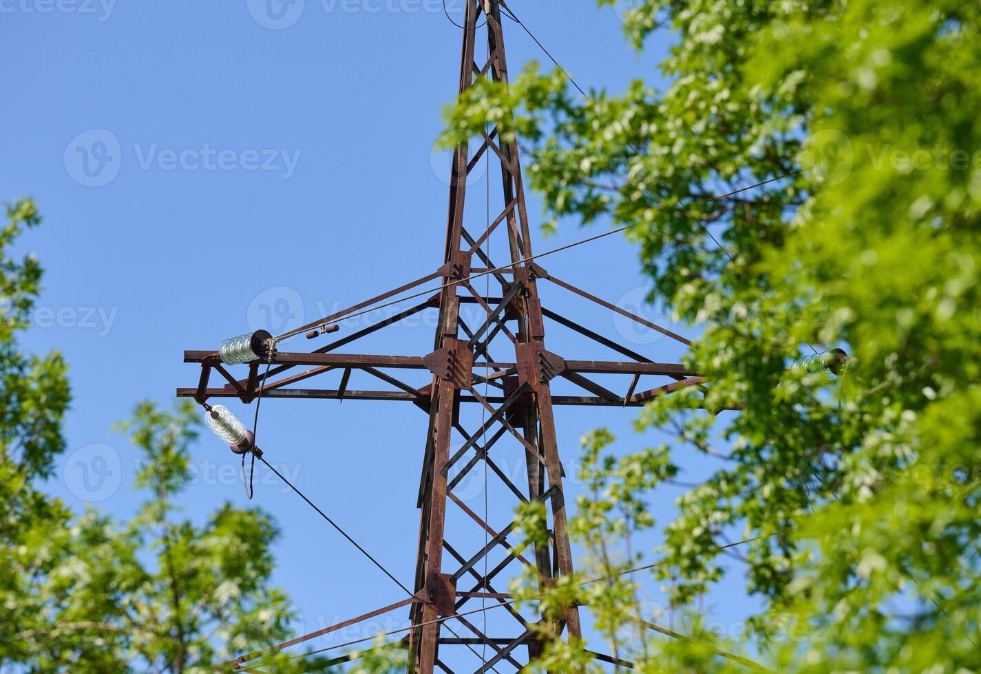 Upper part of an old, rusty power line tower against a blue sky and green tree leaves. photo