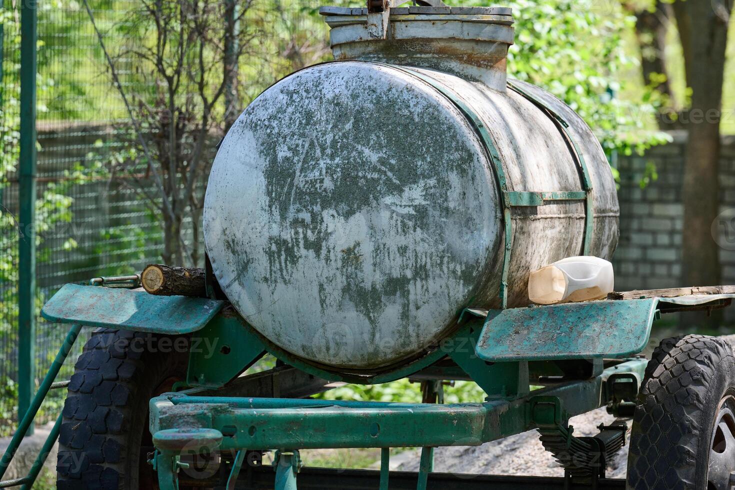 An old metal tank on a trailer, covered with traces of rust and time, stands against the background of green trees and a chain-link fence. photo
