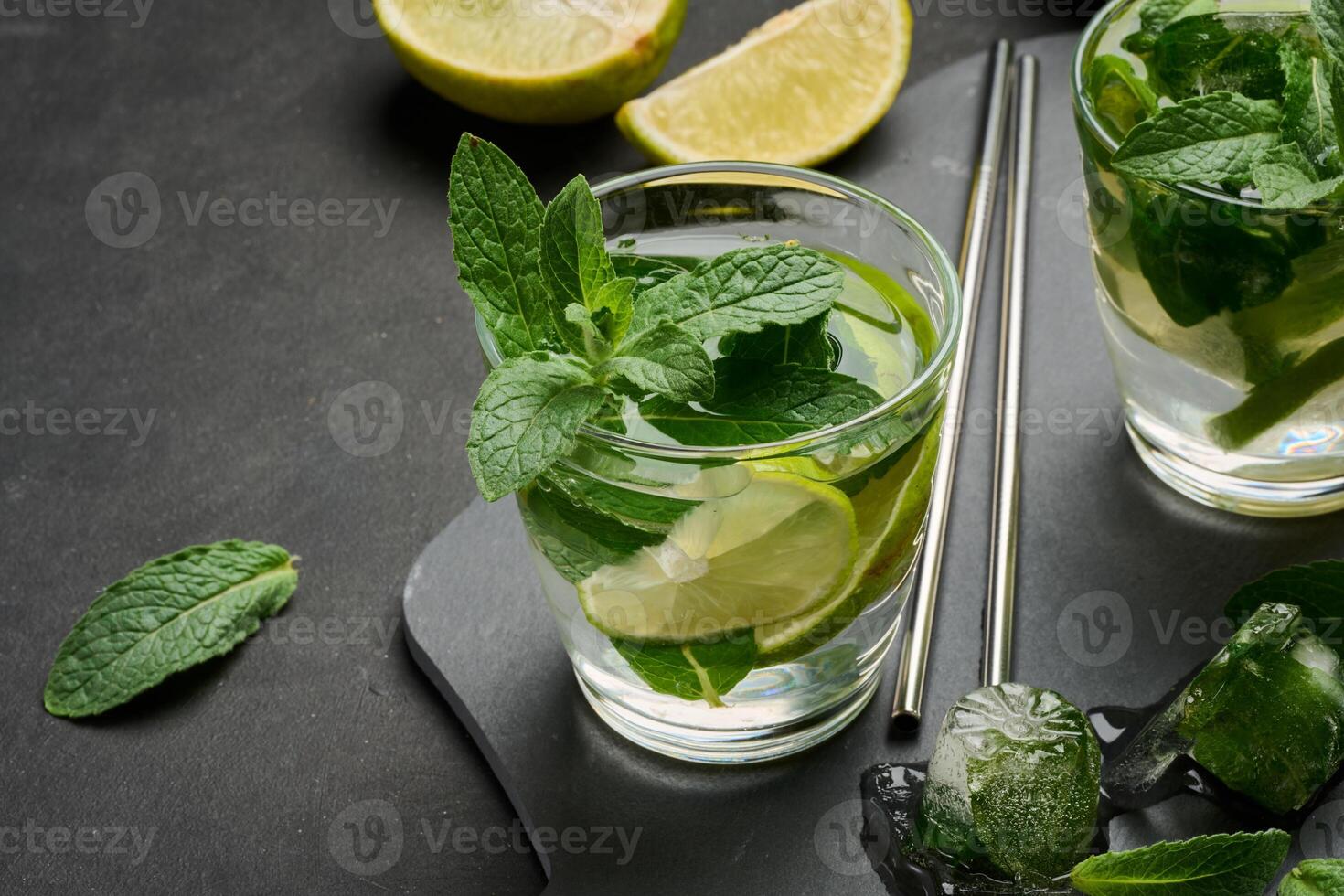 Glass of mojito cocktail with fresh mint leaves, lime slices, and ice cubes on a dark stone background. Refreshing summer drink with stainless steel straws. photo