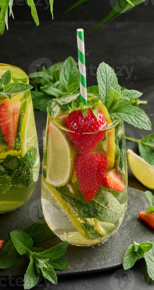 A refreshing drink in a glass with lemon slices, strawberries, fresh mint leaves, and sparkling water, served with lime and mint on a black plate photo