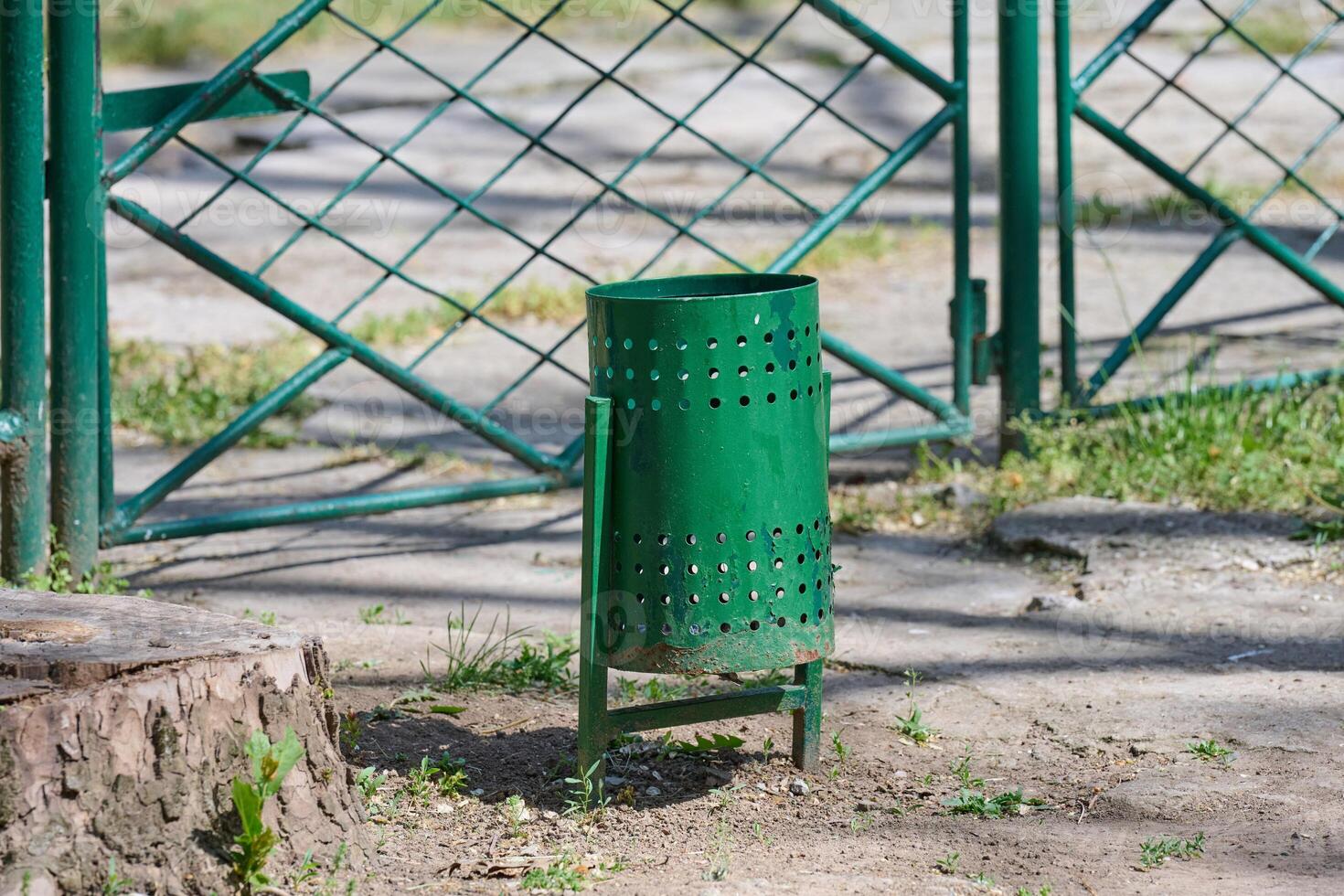 A green perforated metal trash can stands in a park or public area. photo