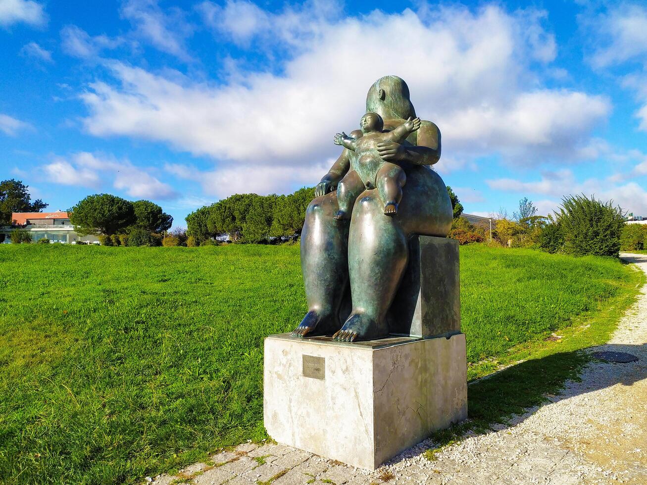 LISBON, PORTUGAL - DECEMBER 04, 2024 Sculpture of mother holding child in public park. Bronze statue against a green lawn and blue sky with clouds. Editorial use. photo