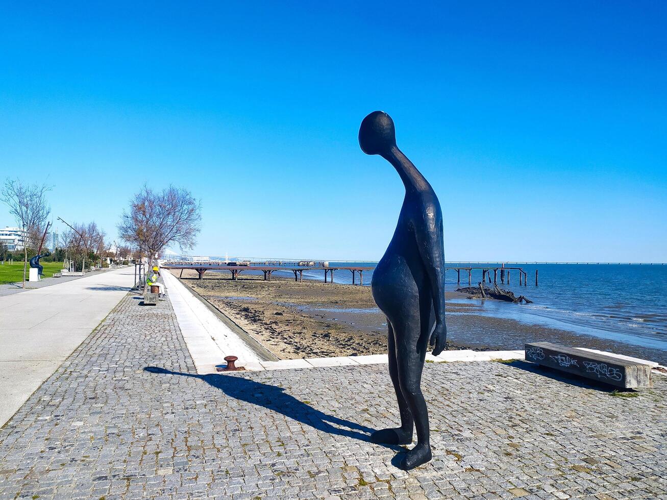 LISBON, PORTUGAL - JANUARY 17, 2025 Modern art sculpture stands on waterfront promenade with bridge and ocean views, showcasing public art and coastal scenery. photo