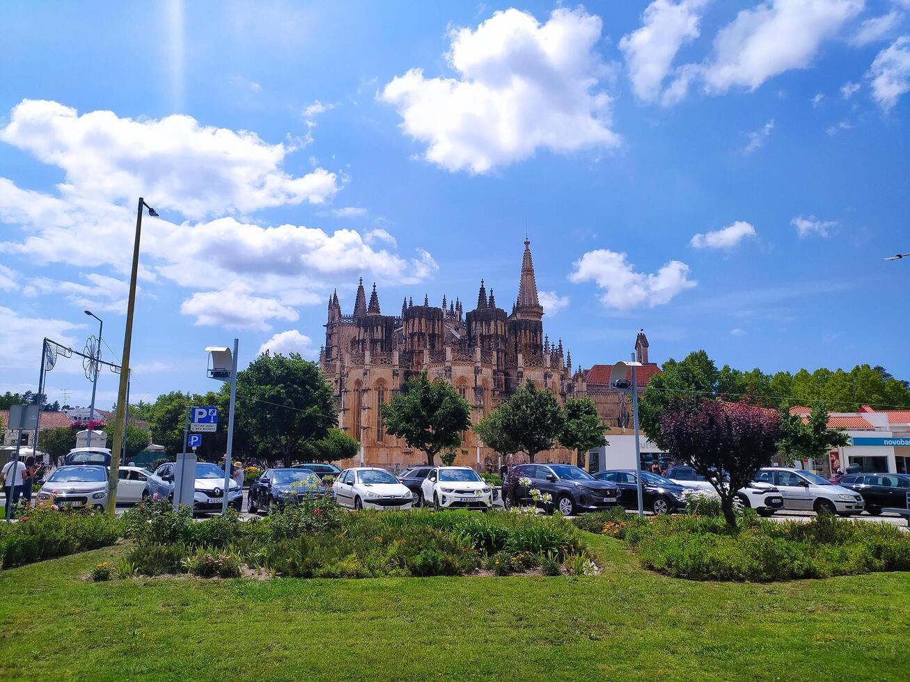 BATALHA, PORTUGAL - JUNE 10, 2023 Scenic view of a historic monastery under a bright sky, parked cars and greenery in foreground. Travel, heritage, architecture. photo