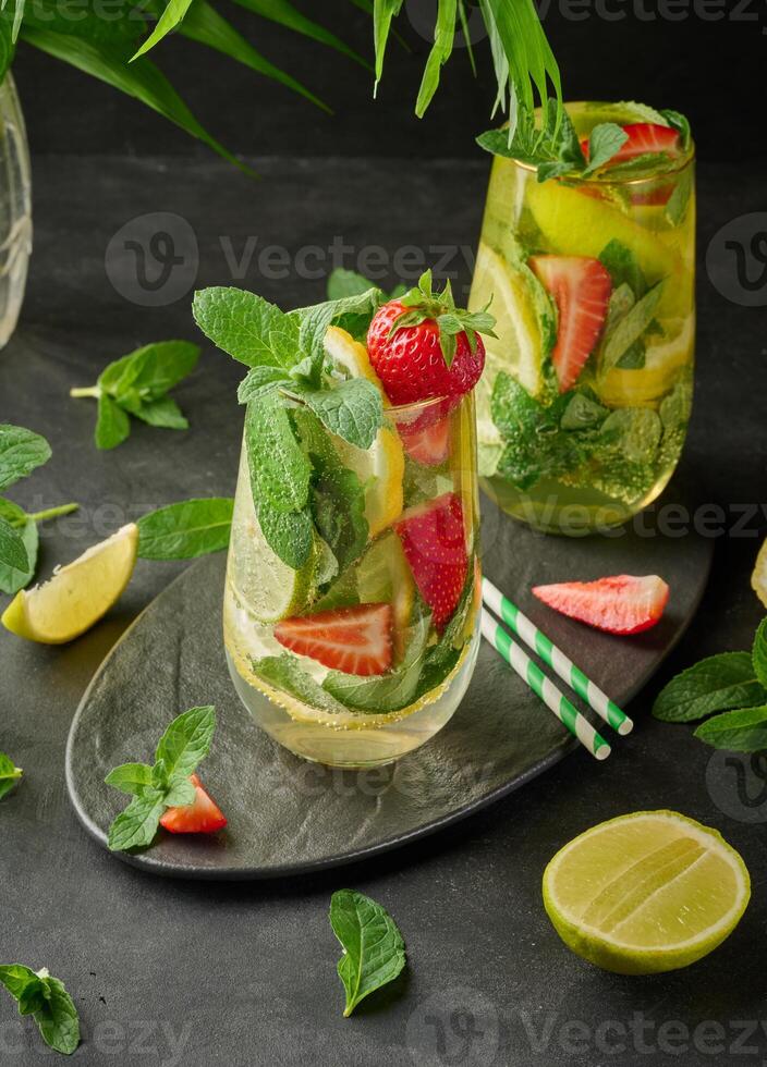 Refreshing drink in a glass with lemon slices, strawberries, fresh mint leaves, and sparkling water, served with lime and mint on a black plate photo