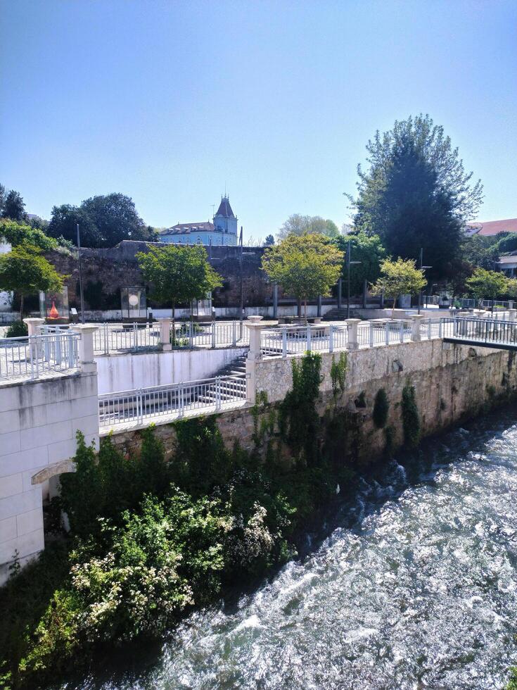 ALCOBACA, PORTUGAL - APRIL 24, 2025 View of a river flowing through a town. Walls, walkways, and greenery are visible. Blue sky and bright daylight. photo