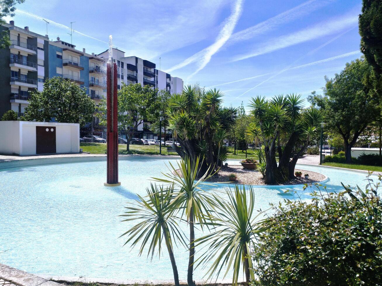 ALCOBACA, PORTUGAL - APRIL 27, 2025 Serene city park with a round water feature, featuring palm trees and green plants, against a backdrop of buildings and a blue sky. photo