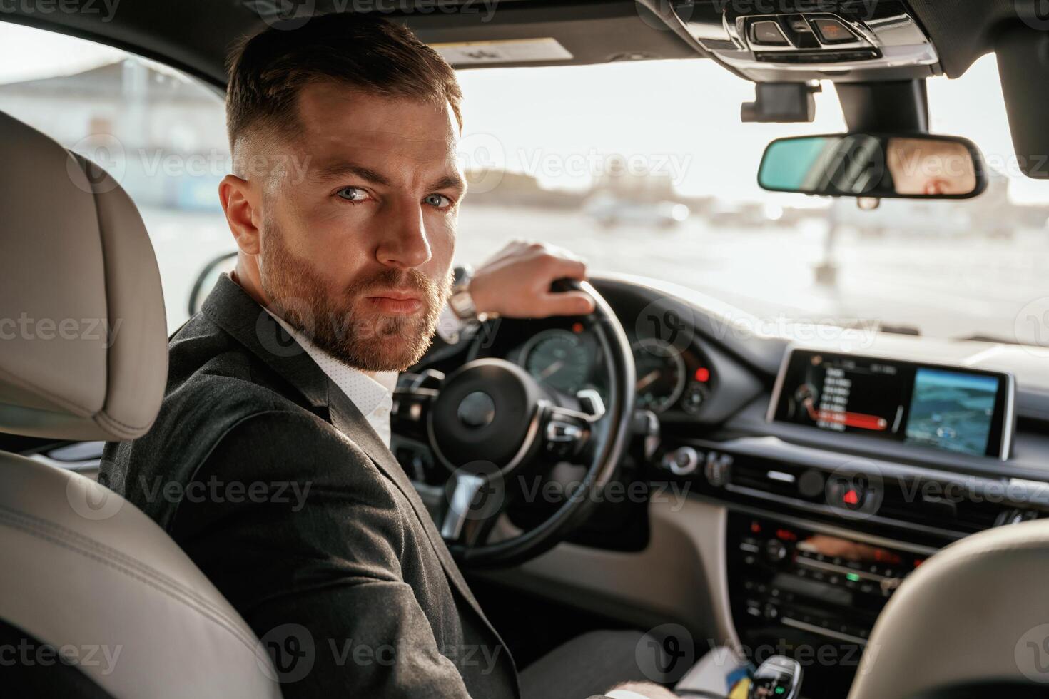 Looking behind at the camera.,, Man ,in ,black suit is sitting in the car, driving photo