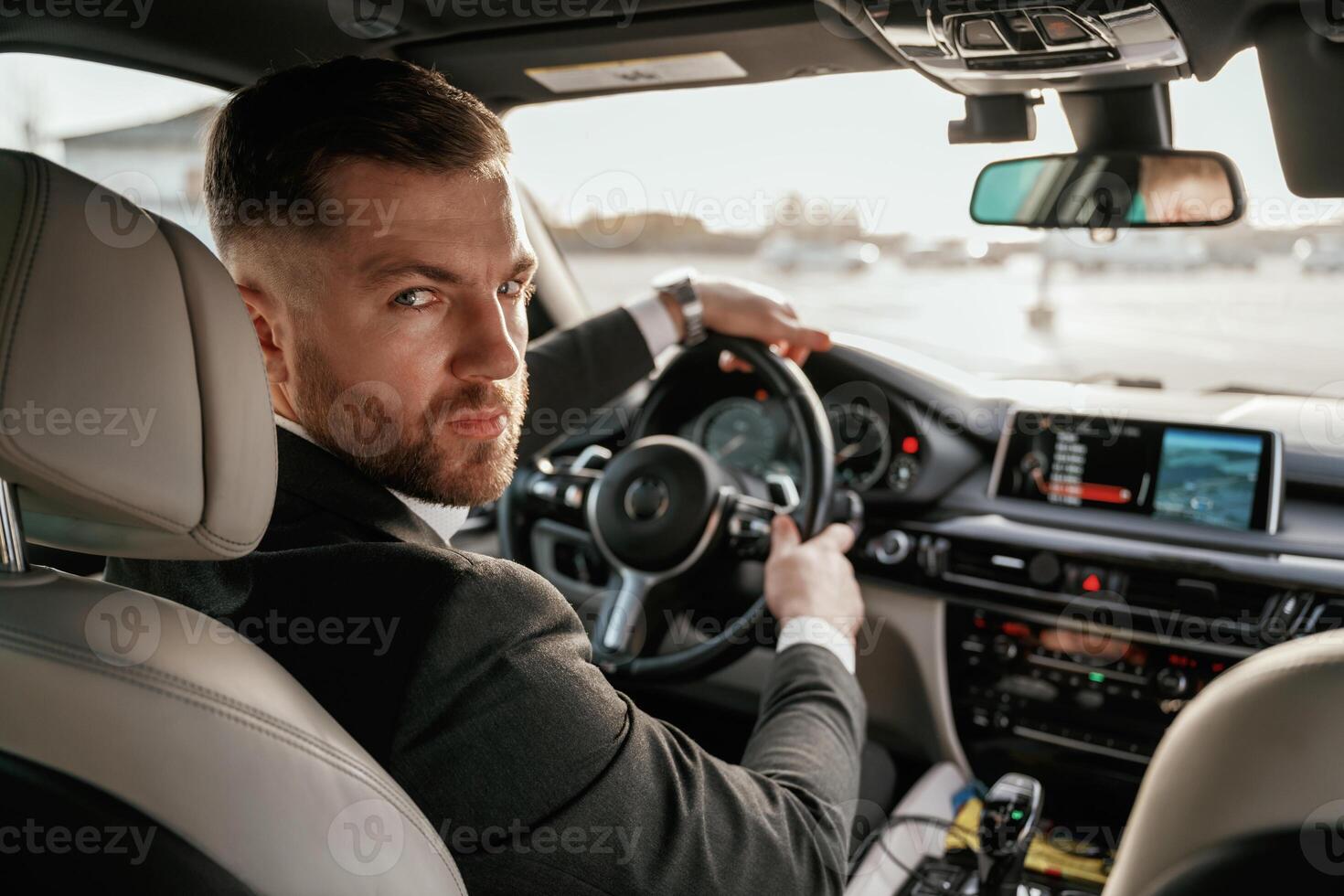 Looking, behind at the camera. Man in black suit is sitting in the car, driving photo