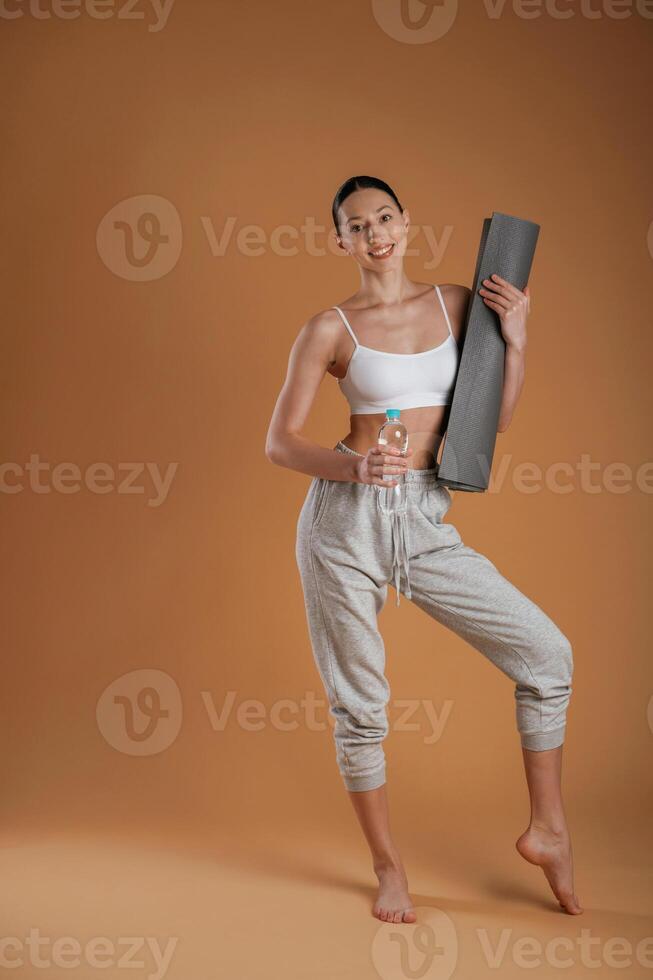 Looking forward, holding bottle of water and mat. Beautiful fitness woman is in the studio photo
