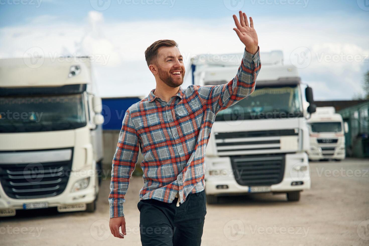 Greetings, hello hand gesture, smiling. Man is standing against trucks outdoors photo