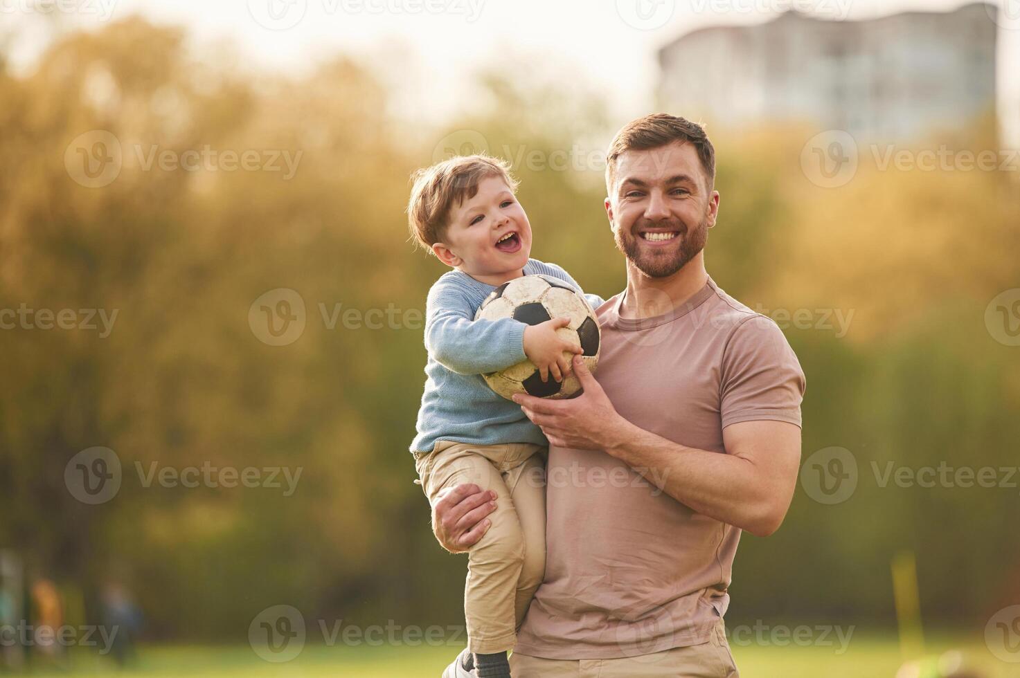 Standing, holding soccer ball. Happy father with son are having fun on the field at summertime photo