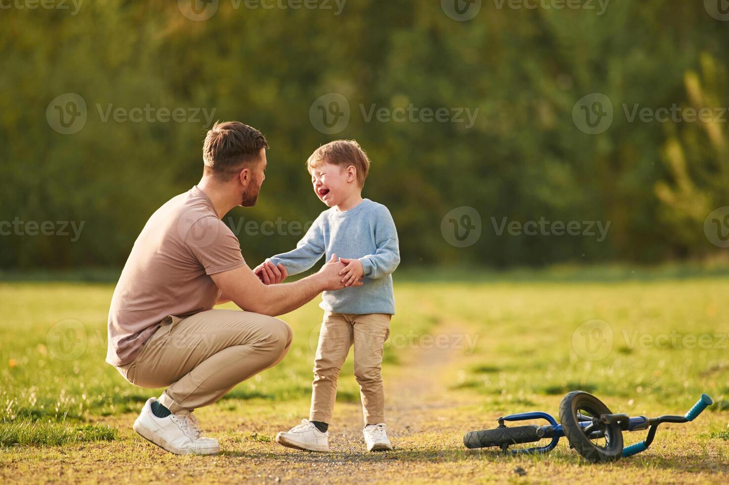 Boy is crying, fell of the bicycle. Happy father with son are having fun on the field at summertime photo