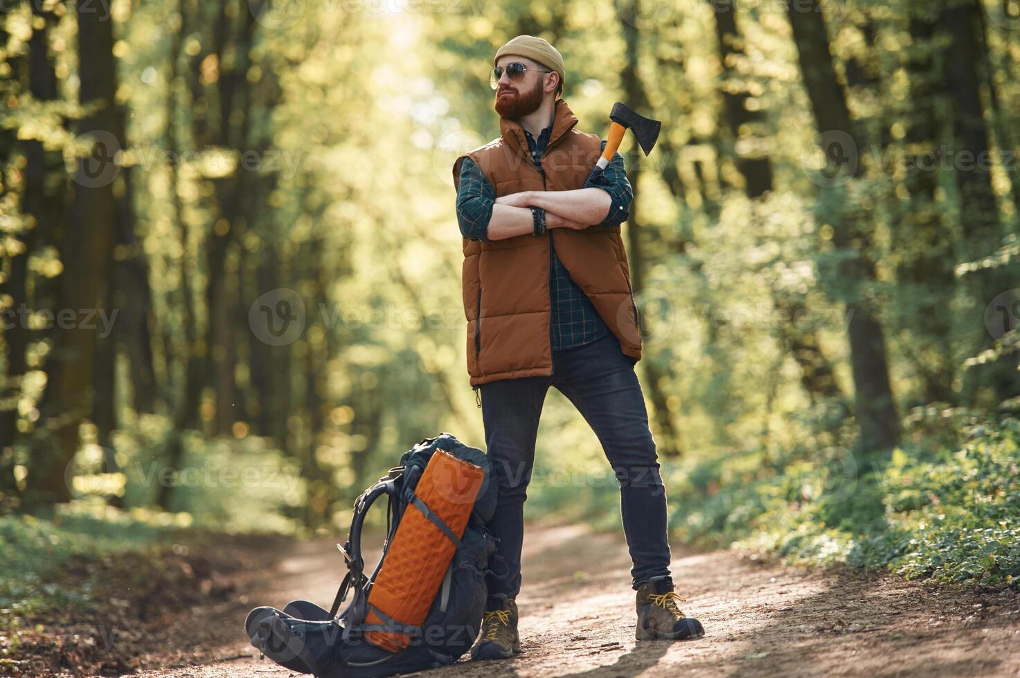 Yellow, axe in ,hand, backpack on the ground. Bearded man is in the forest at daytime photo