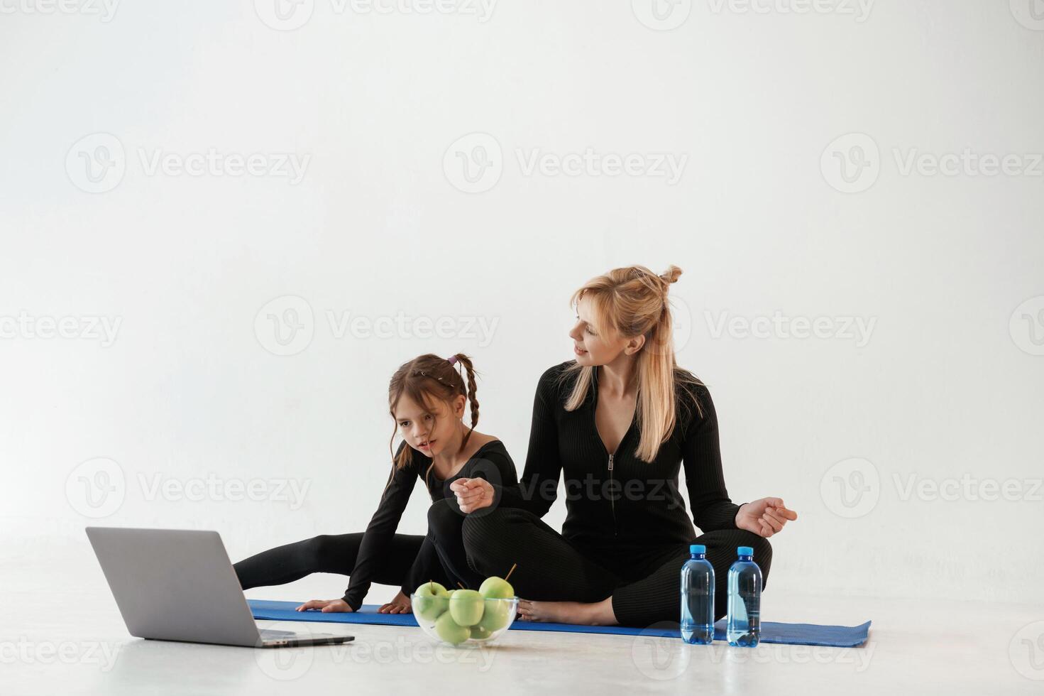 Laptop, apples ,and bottles of water on the floor. Mother and little daughter are in fitness clothes on yoga mat photo