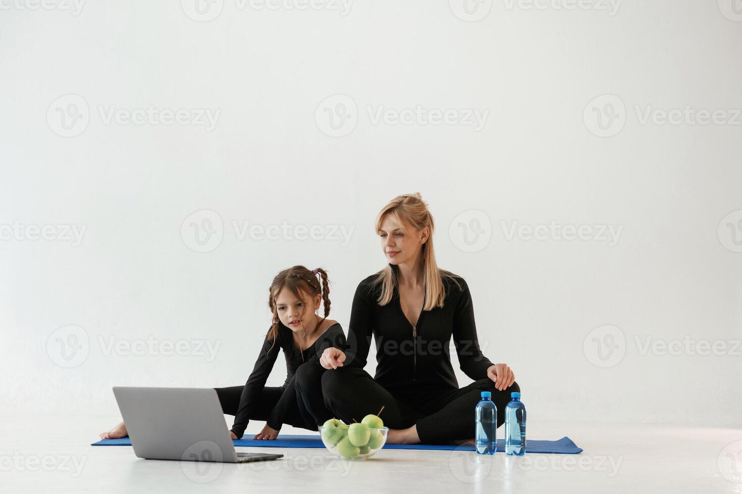 Laptop,,, apples and bottles of water on the floor. Mother and little daughter are in fitness clothes on yoga mat photo