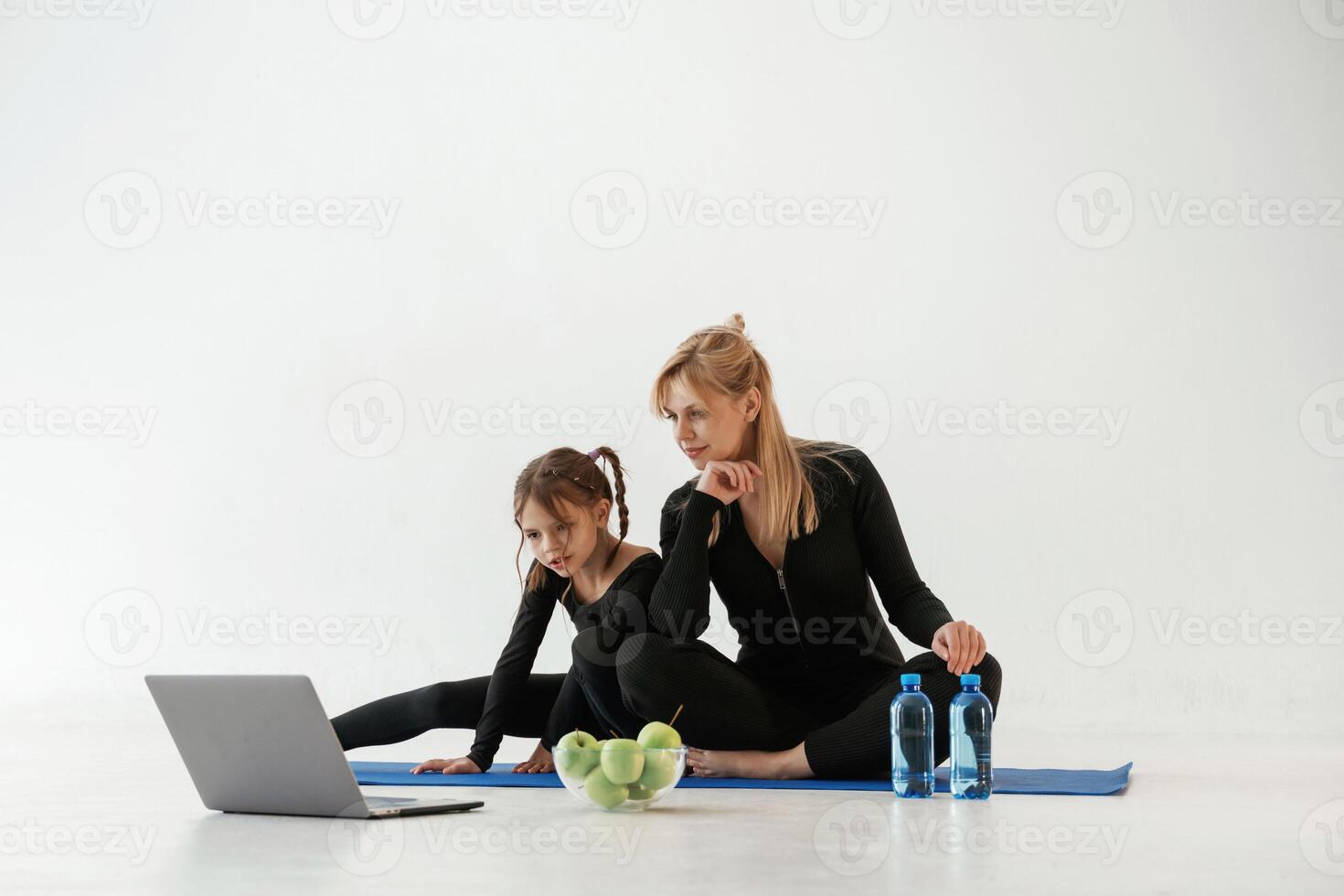 Laptop,, apples and bottles of water on the floor. Mother and little daughter are in fitness clothes on yoga mat photo