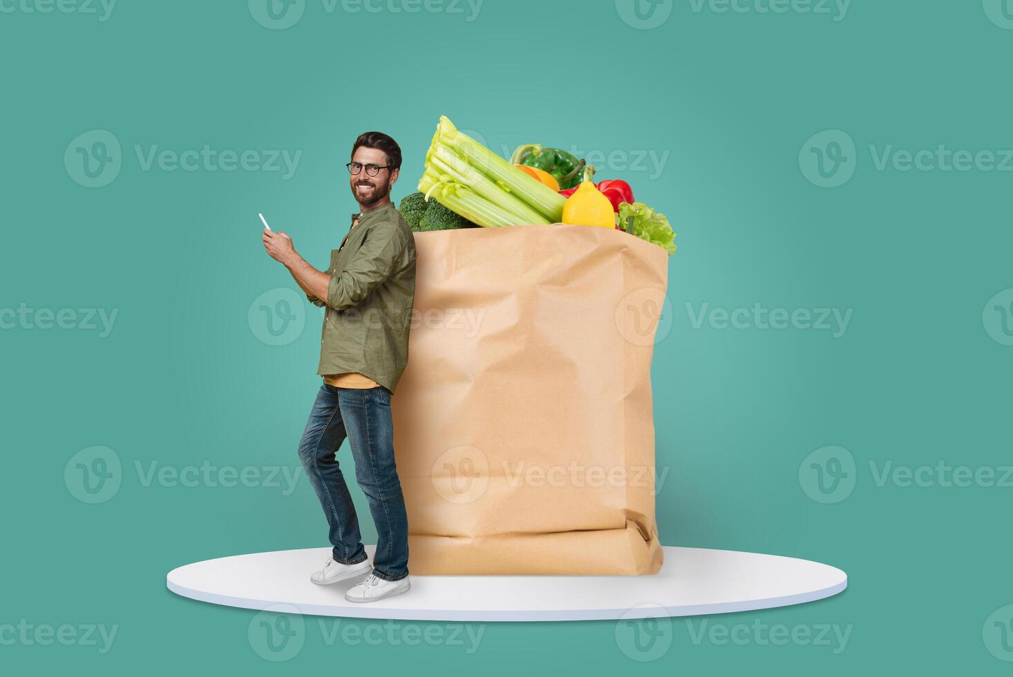 A cheerful man stands next to a large paper bag filled with fresh vegetables, showcasing the new normal of grocery shopping and delivery while checking his phone. photo