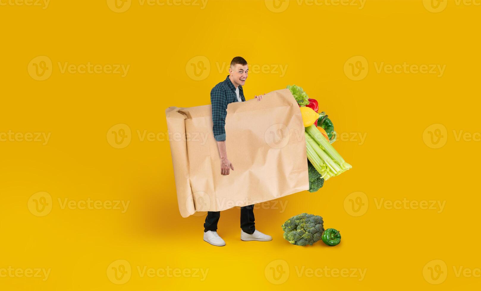 A person is smiling while holding a large paper bag overflowing with various groceries, including fresh vegetables. The vibrant yellow background enhances the cheerful atmosphere of grocery shopping. photo