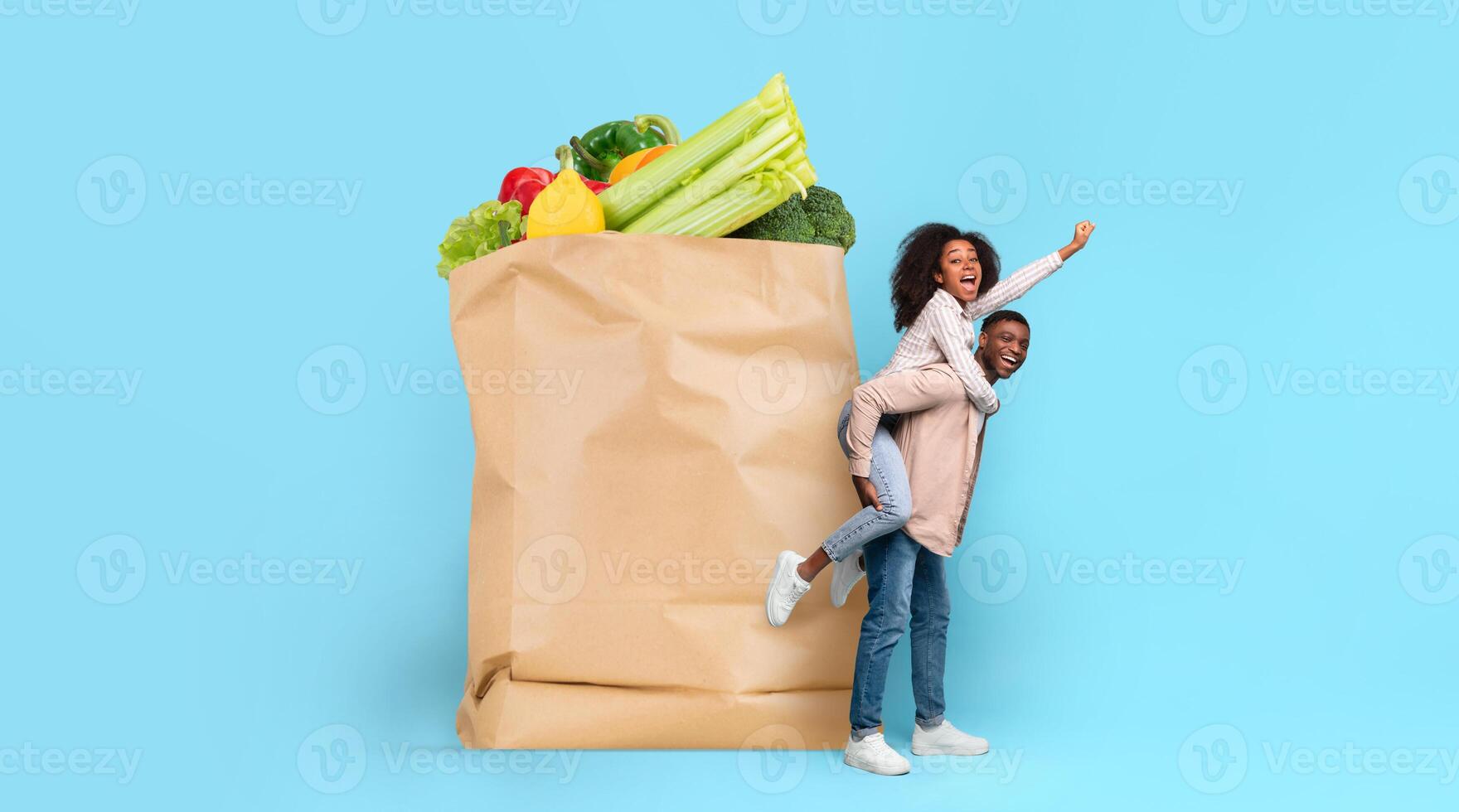 A couple is playfully engaged in grocery shopping, with one person on the other's back. Fresh vegetables and fruits are visible in a large paper bag, symbolizing the joys of cooking together. photo