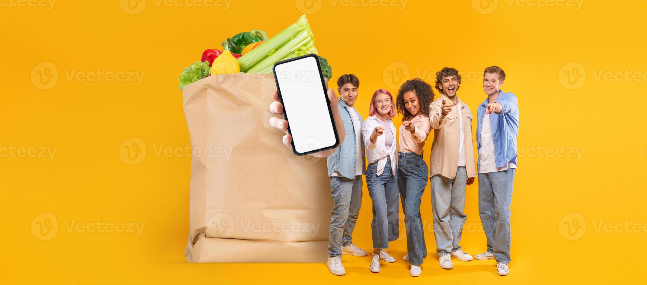 Five individuals smile while showcasing a smartphone in front of a paper grocery bag filled with fresh produce. This reflects modern shopping trends in an isolated background. photo
