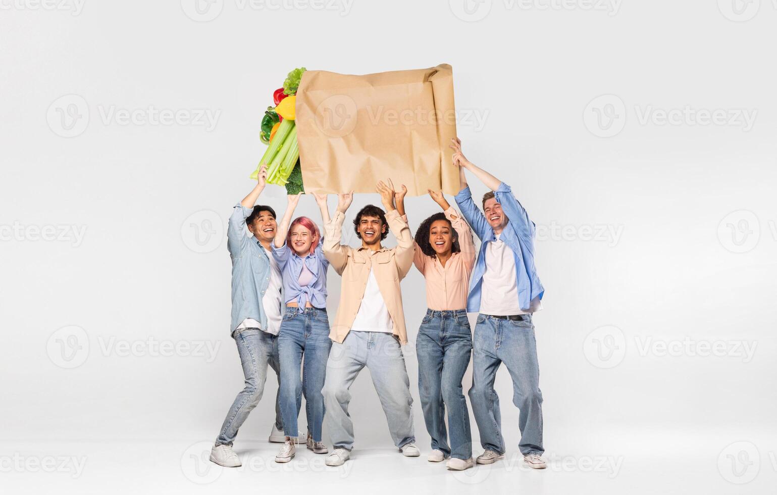 A group of five friends joyfully holds up a large paper bag filled with groceries. photo