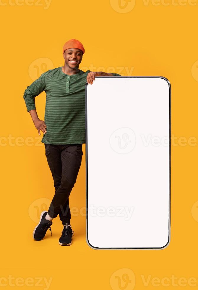 Mobile Offer. Happy Black Guy Leaning At Big Smartphone With Blank White Screen, Cheerful African Man Demonstrating Copy Space For Your Design Or Advertisement, Posing Over Yellow Background, Mockup photo
