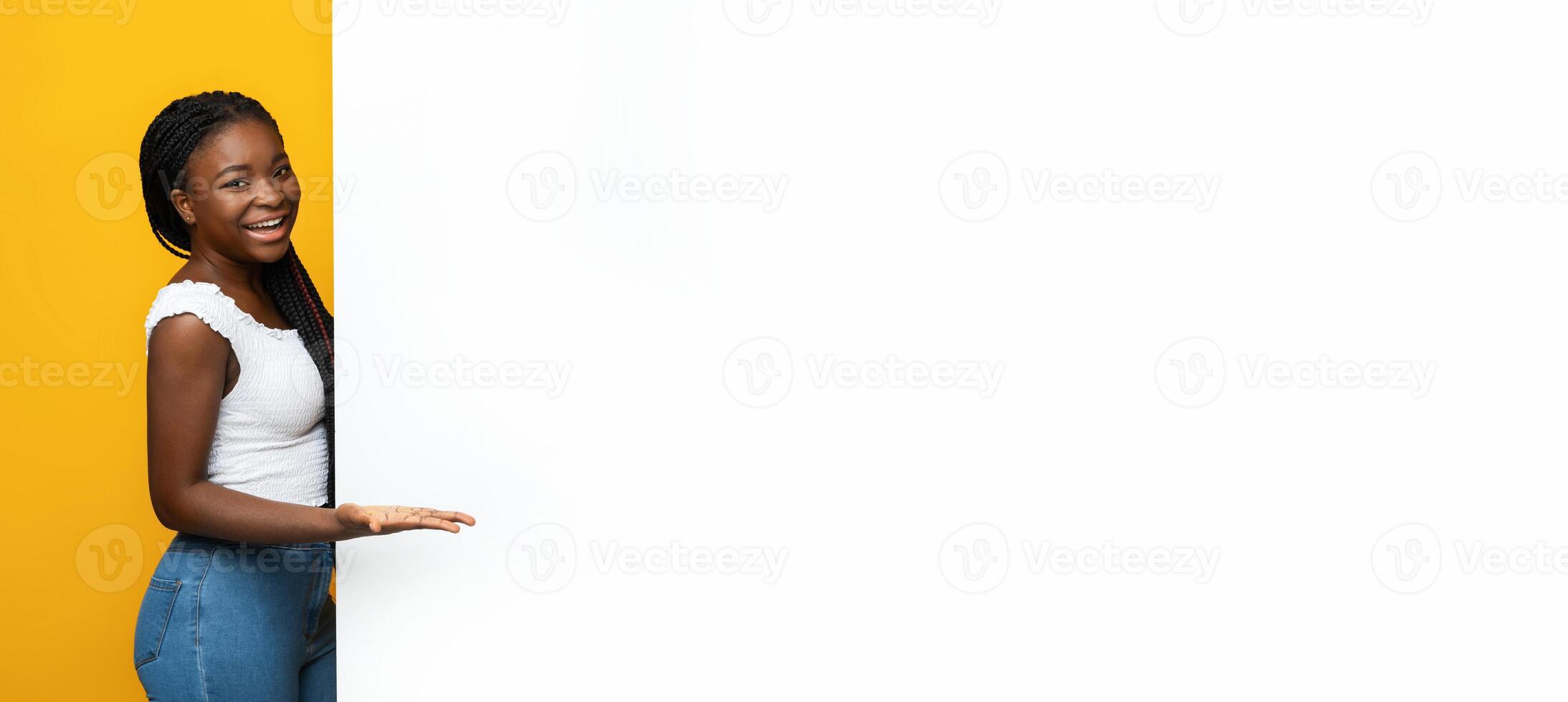 Cheerful black woman pointing at white advertisement board with palm, demonstrating copy space for your text or design, standing near blank billboard over yellow studio background, panorama photo