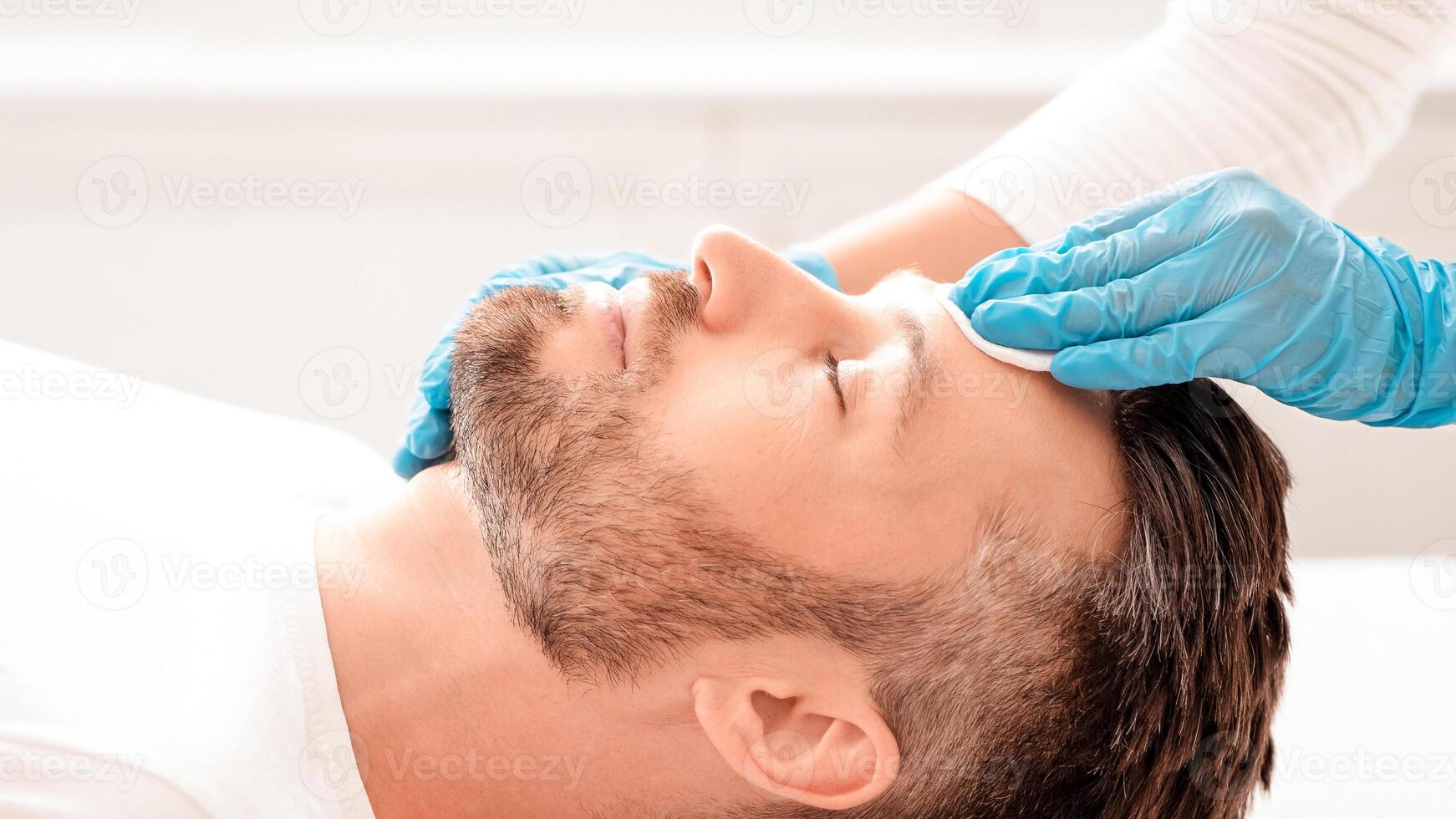 Side view of unrecognizable cosmetologist cleansing bearded man face with toner before or after treatment, using cotton pad. Middle-aged businessman having beauty procedures at spa photo