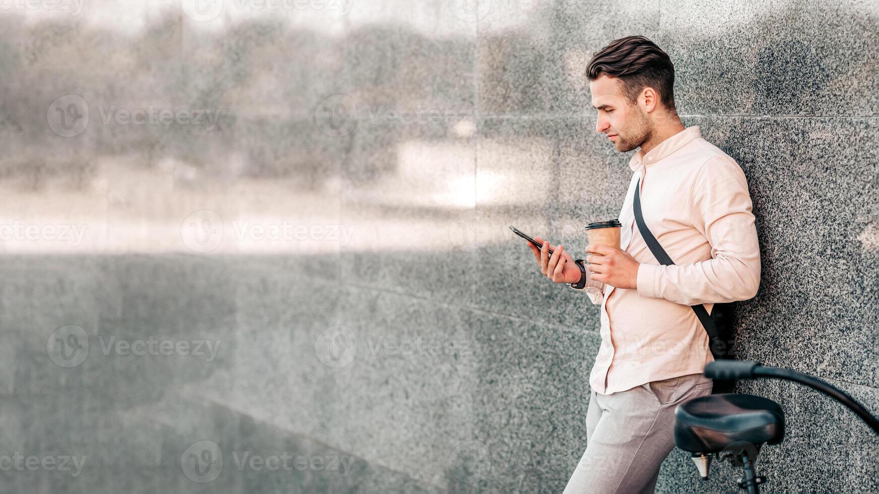 A young man leans against a sleek wall on a city street, sipping coffee and checking his phone. Dressed casually, he embodies a modern lifestyle while surrounded by urban scenery, copy space photo