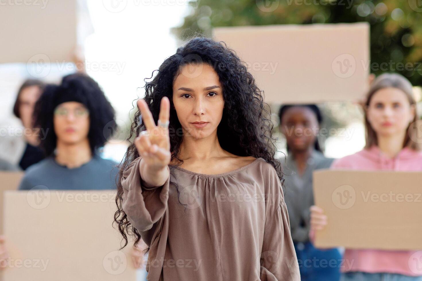 Multiracial group of demonstrators with blank placards fighting for peace in the world, walking by street. Mixed race young woman leading group of international protestors, showing peace gesture photo